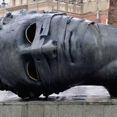 Mroczna, wielka rzeźba głowy Igora Mitoraja Eros Bendato z pustymi oczodołami, na tle zabytkowej architektury.