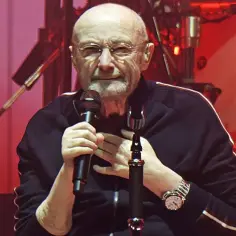 Phil Collins, mimo choroby, trzyma mikrofon i opowiada publiczności.